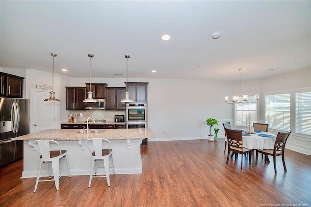 kitchen with a kitchen island with sink, stainless steel appliances, wood finished floors, dark brown cabinets, and decorative backsplash