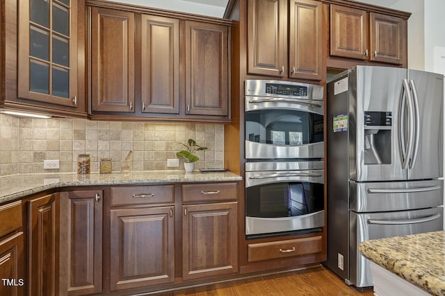 kitchen featuring tasteful backsplash, glass insert cabinets, light stone countertops, wood finished floors, and stainless steel appliances