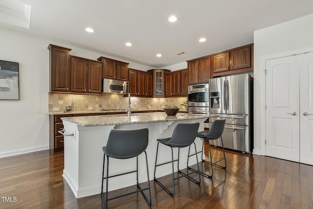 kitchen with backsplash, light stone countertops, dark wood-style flooring, appliances with stainless steel finishes, and a kitchen island with sink