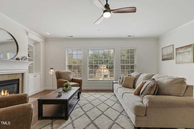 living room with built in shelves, visible vents, light wood-style floors, a glass covered fireplace, and crown molding