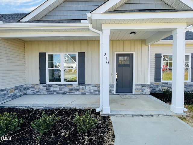 view of exterior entry with stone siding, covered porch, and roof with shingles