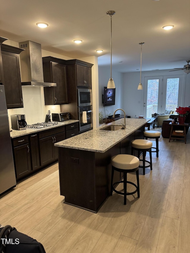 kitchen with light wood-style floors, stainless steel appliances, wall chimney range hood, a kitchen bar, and a sink