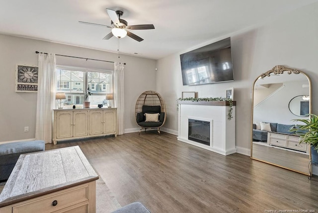 living area with a glass covered fireplace, wood finished floors, a ceiling fan, and baseboards