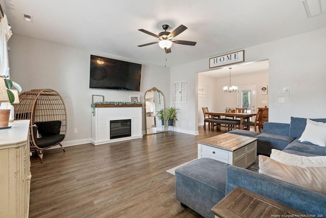 living area with dark wood-style floors, visible vents, a glass covered fireplace, baseboards, and ceiling fan with notable chandelier