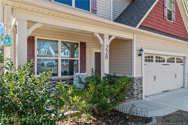 doorway to property featuring roof with shingles, concrete driveway, covered porch, a garage, and stone siding