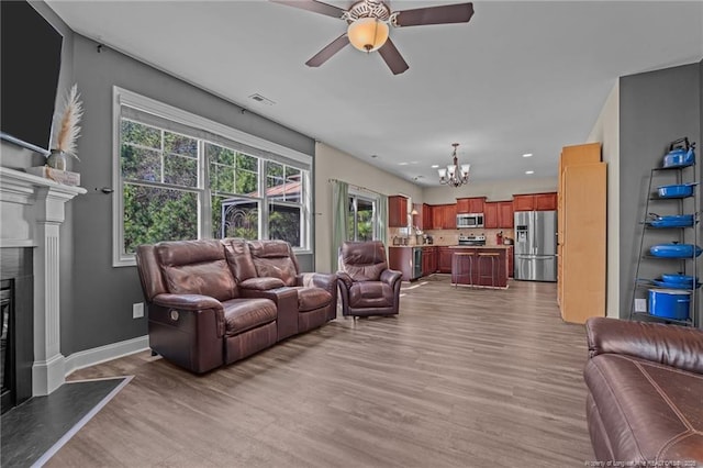 living room with light wood-type flooring, a glass covered fireplace, baseboards, and ceiling fan with notable chandelier