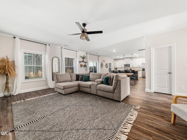 living room with dark wood-type flooring, crown molding, baseboards, and ceiling fan