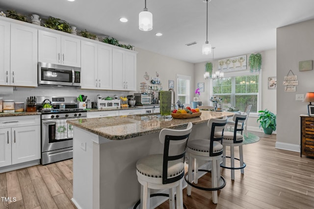 kitchen featuring light wood-style flooring, stainless steel appliances, backsplash, dark stone counters, and an island with sink