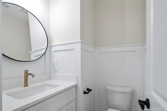 half bath with toilet, a decorative wall, vanity, and wainscoting