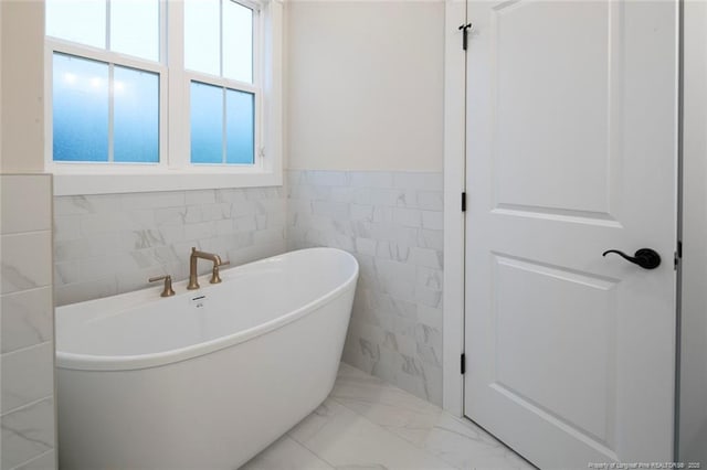 full bathroom featuring a soaking tub, marble finish floor, wainscoting, and tile walls
