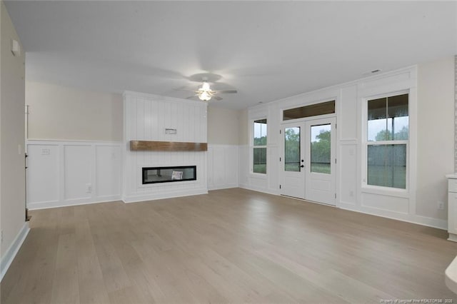 unfurnished living room with a large fireplace, a ceiling fan, light wood-style floors, wainscoting, and a decorative wall