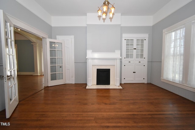 unfurnished living room featuring arched walkways, wainscoting, a fireplace with flush hearth, dark wood-style flooring, and ornate columns