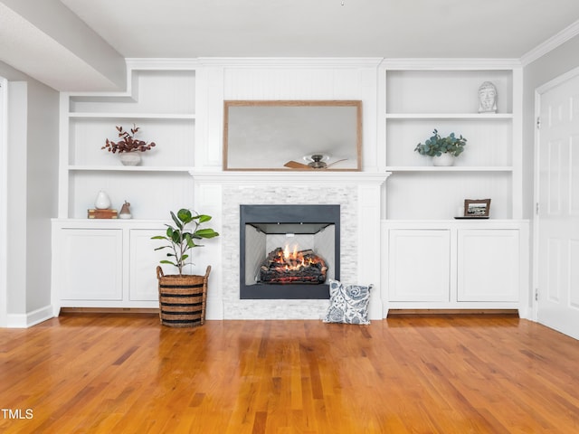 unfurnished living room with built in features, a ceiling fan, ornamental molding, wood finished floors, and a lit fireplace