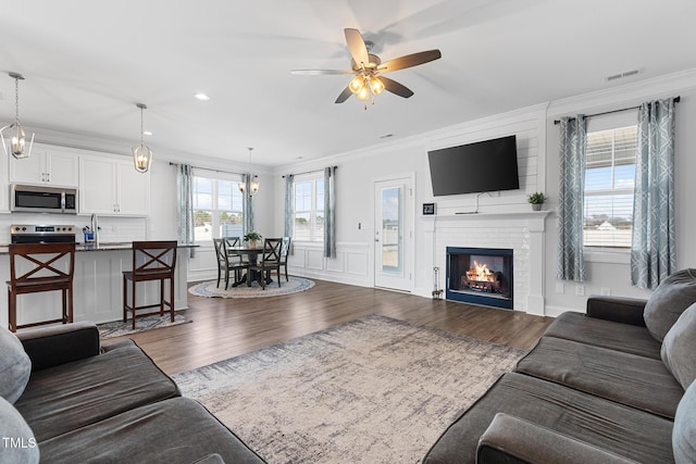 living room with dark wood finished floors, crown molding, a fireplace, a decorative wall, and ceiling fan with notable chandelier