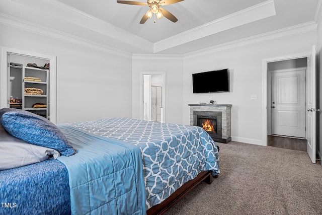 carpeted bedroom with a warm lit fireplace, baseboards, a raised ceiling, a ceiling fan, and crown molding