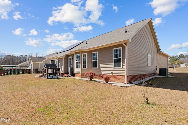 rear view of house with cooling unit, a shingled roof, fence, a lawn, and roof mounted solar panels