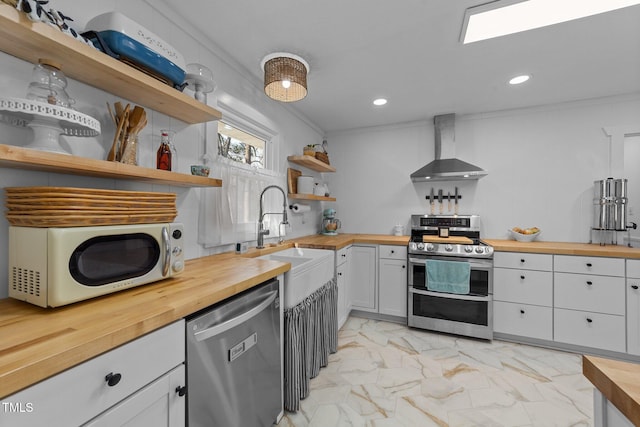 kitchen with open shelves, butcher block countertops, stainless steel appliances, a sink, and wall chimney exhaust hood