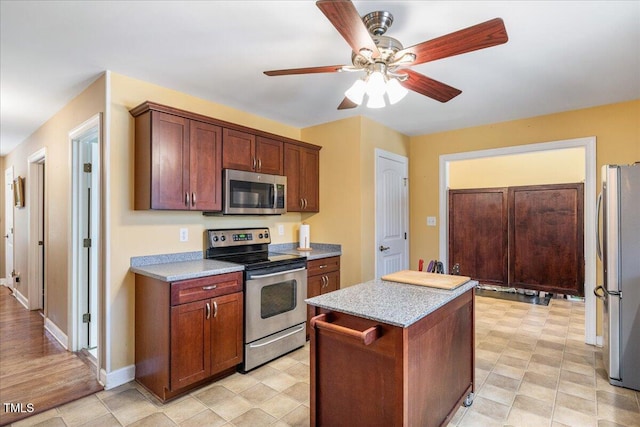 kitchen with ceiling fan, light stone counters, stainless steel appliances, a kitchen island, and baseboards