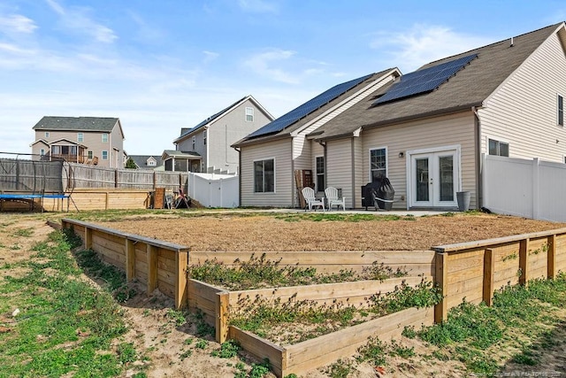 rear view of property featuring a fenced backyard, french doors, a vegetable garden, roof mounted solar panels, and a trampoline