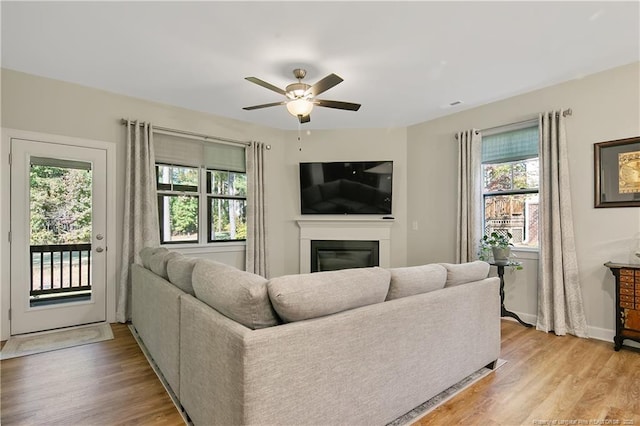 living area with light wood-style floors, baseboards, a ceiling fan, and a glass covered fireplace