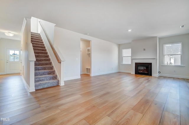 unfurnished living room with light wood-style flooring, stairway, baseboards, and a glass covered fireplace