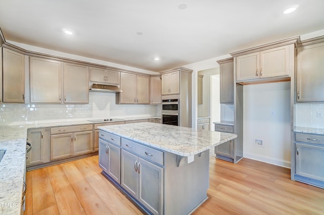 kitchen with light wood finished floors, tasteful backsplash, a kitchen island, light stone countertops, and under cabinet range hood