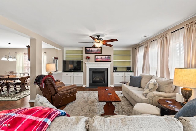 living room featuring dark wood-style floors, a fireplace with flush hearth, visible vents, and ceiling fan with notable chandelier