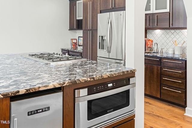 kitchen featuring tasteful backsplash, light wood-style flooring, appliances with stainless steel finishes, dark brown cabinets, and dark stone counters