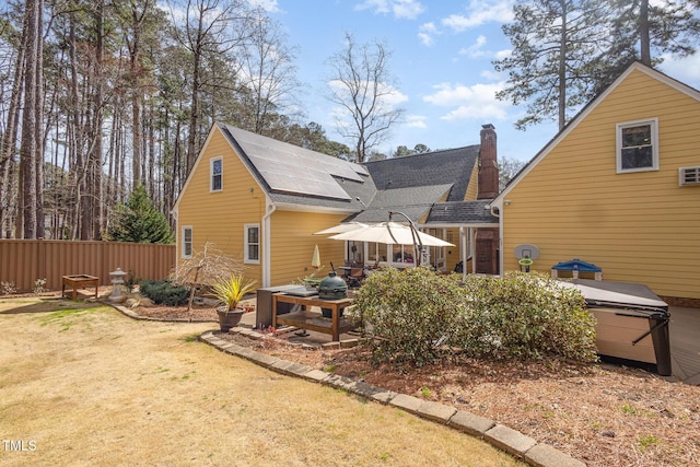 back of house featuring a jacuzzi, fence, roof with shingles, roof mounted solar panels, and a chimney