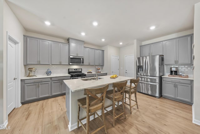 kitchen featuring appliances with stainless steel finishes, a kitchen island with sink, gray cabinets, light wood-type flooring, and a sink