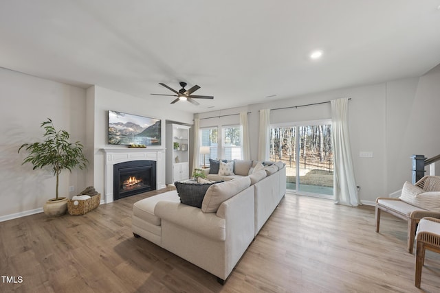 living area featuring baseboards, built in features, a glass covered fireplace, ceiling fan, and light wood-type flooring