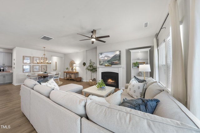 living room featuring light wood-style floors, a lit fireplace, visible vents, and ceiling fan with notable chandelier