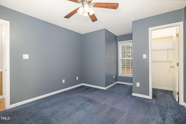 unfurnished bedroom with a textured ceiling, a ceiling fan, baseboards, a spacious closet, and dark colored carpet