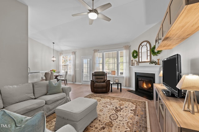 living area featuring baseboards, ceiling fan, light wood-style flooring, a fireplace with flush hearth, and vaulted ceiling