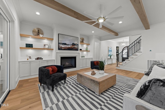 living room featuring stairs, a lit fireplace, light wood finished floors, and beamed ceiling