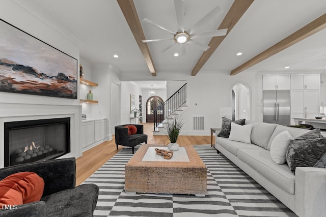 living area featuring beam ceiling, visible vents, light wood-style floors, a lit fireplace, and stairs