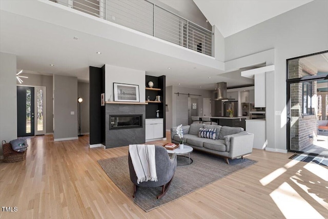 living room featuring light wood finished floors, baseboards, a tiled fireplace, a high ceiling, and a ceiling fan