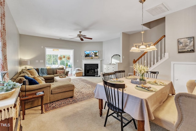 dining area with light colored carpet, ceiling fan with notable chandelier, visible vents, stairway, and a glass covered fireplace