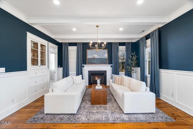 living area featuring an inviting chandelier, wainscoting, wood finished floors, beamed ceiling, and a lit fireplace