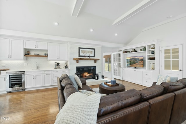 living area with beverage cooler, wet bar, vaulted ceiling with beams, light wood-style floors, and a brick fireplace