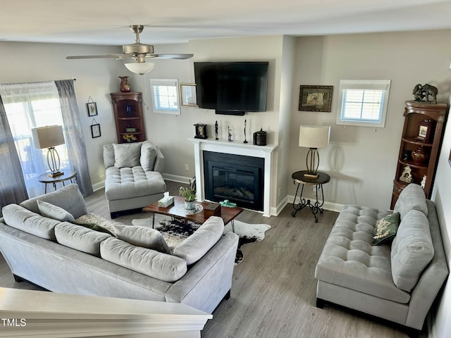 living area with a glass covered fireplace, ceiling fan, baseboards, and wood finished floors