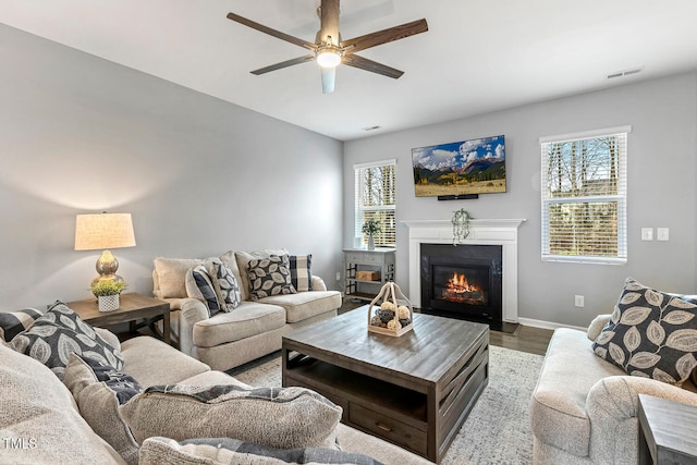 living room featuring visible vents, a ceiling fan, wood finished floors, a glass covered fireplace, and baseboards