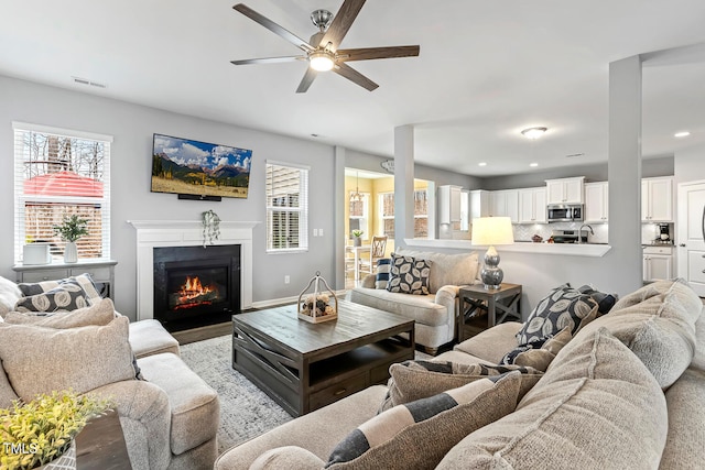 living area featuring visible vents, plenty of natural light, a ceiling fan, and a glass covered fireplace