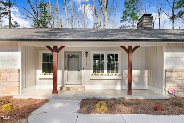 property entrance with stone siding, a chimney, covered porch, and roof with shingles