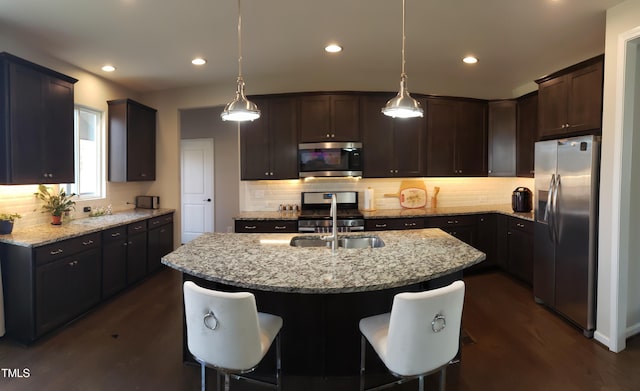 kitchen with light stone counters, an island with sink, stainless steel appliances, dark brown cabinetry, and dark wood-type flooring