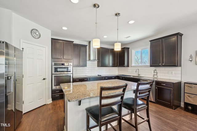 kitchen with dark brown cabinets, a center island, appliances with stainless steel finishes, wall chimney exhaust hood, and dark wood-style flooring