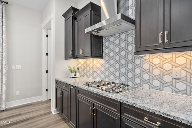 kitchen featuring light stone counters, stainless steel gas cooktop, wall chimney exhaust hood, tasteful backsplash, and baseboards