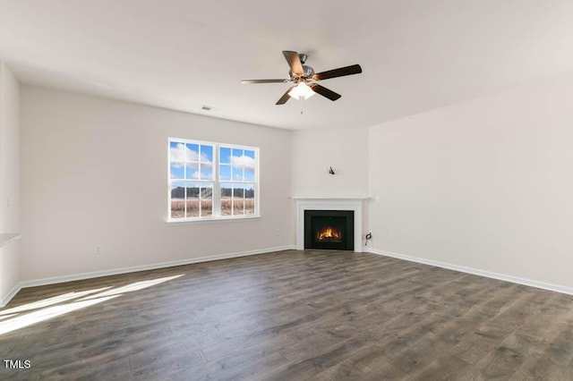 unfurnished living room with a lit fireplace, baseboards, ceiling fan, and dark wood-style flooring