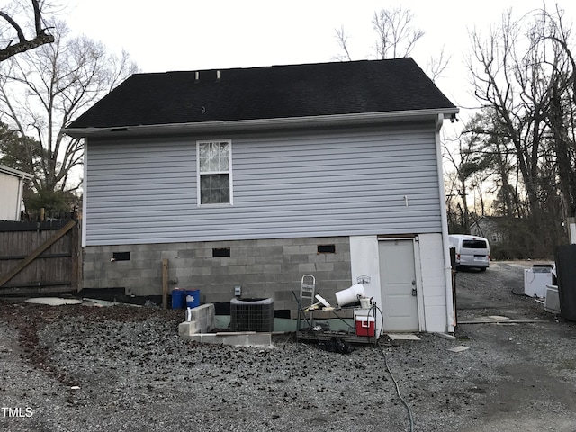 rear view of house with central AC unit, a shingled roof, fence, driveway, and crawl space