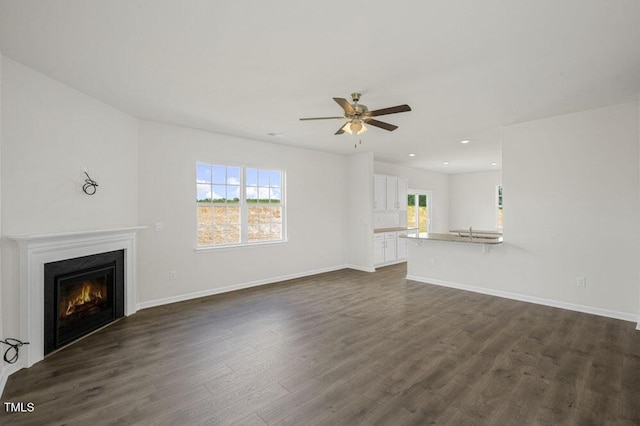 unfurnished living room with a warm lit fireplace, baseboards, ceiling fan, dark wood-style flooring, and recessed lighting